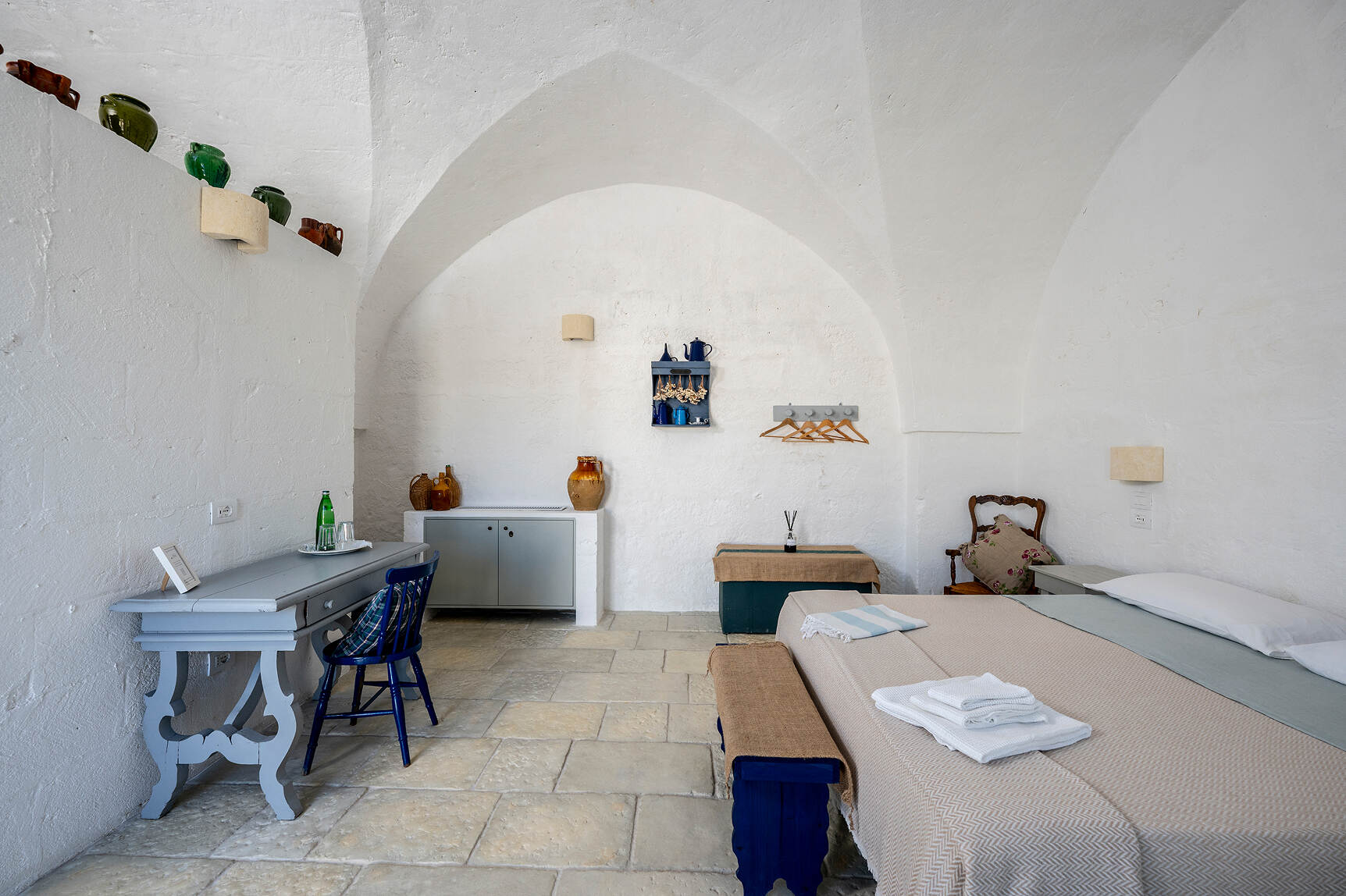 Bright airy bedroom with white vaulted ceilings, stone tile floor, beige bedspread on double bed with folded towels, blue bench at foot, pale blue desk with blue chair, wooden chair with floral cushion, and shelves holding ceramic pots and hanging coat hangers.