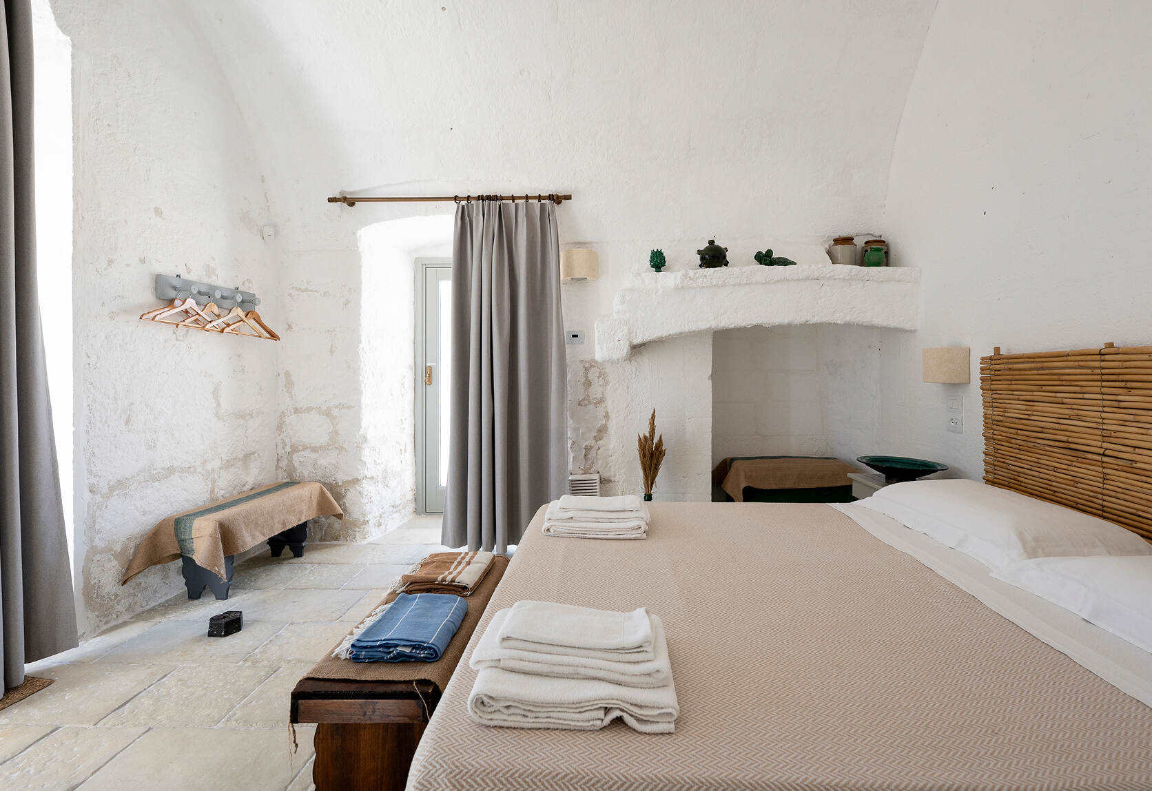 Bright minimalist bedroom with white textured walls, a beige bed with white pillows and folded towels on top, a wooden bench at the foot of the bed with folded blankets, a small table with decor inside a white alcove, and a window with gray curtains.