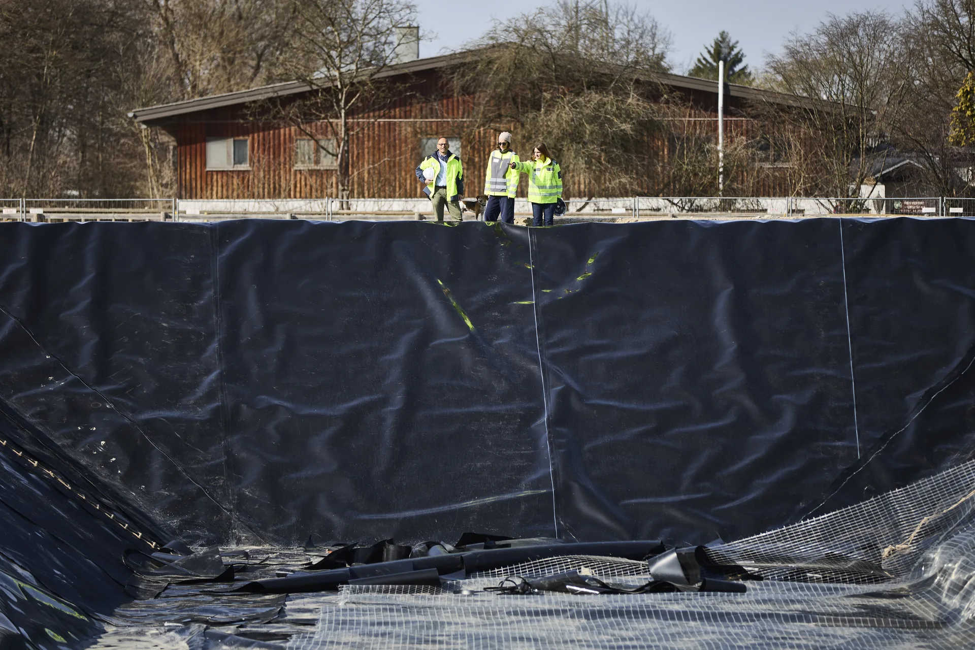 Blick in das mit dunkler Dichtungsbahn ausgekleidete Testwasserbecken; drei Personen stehen am oberen Rand.