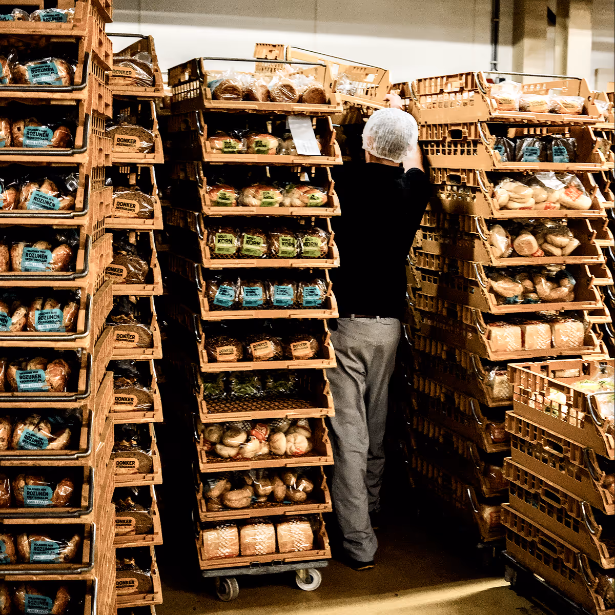 Werknemer met haarnet plaatst dozen met verpakt brood op stapels in een magazijn.