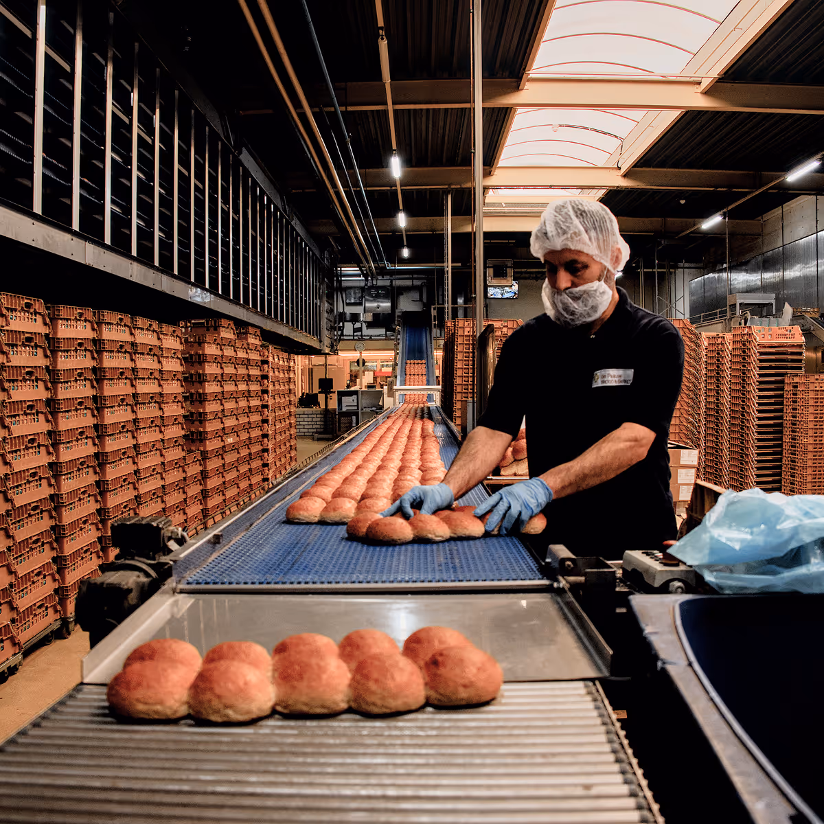Werknemer met handschoenen, haarnetje en mondkapje sorteert broodjes op een lopende band in een bakkerij.