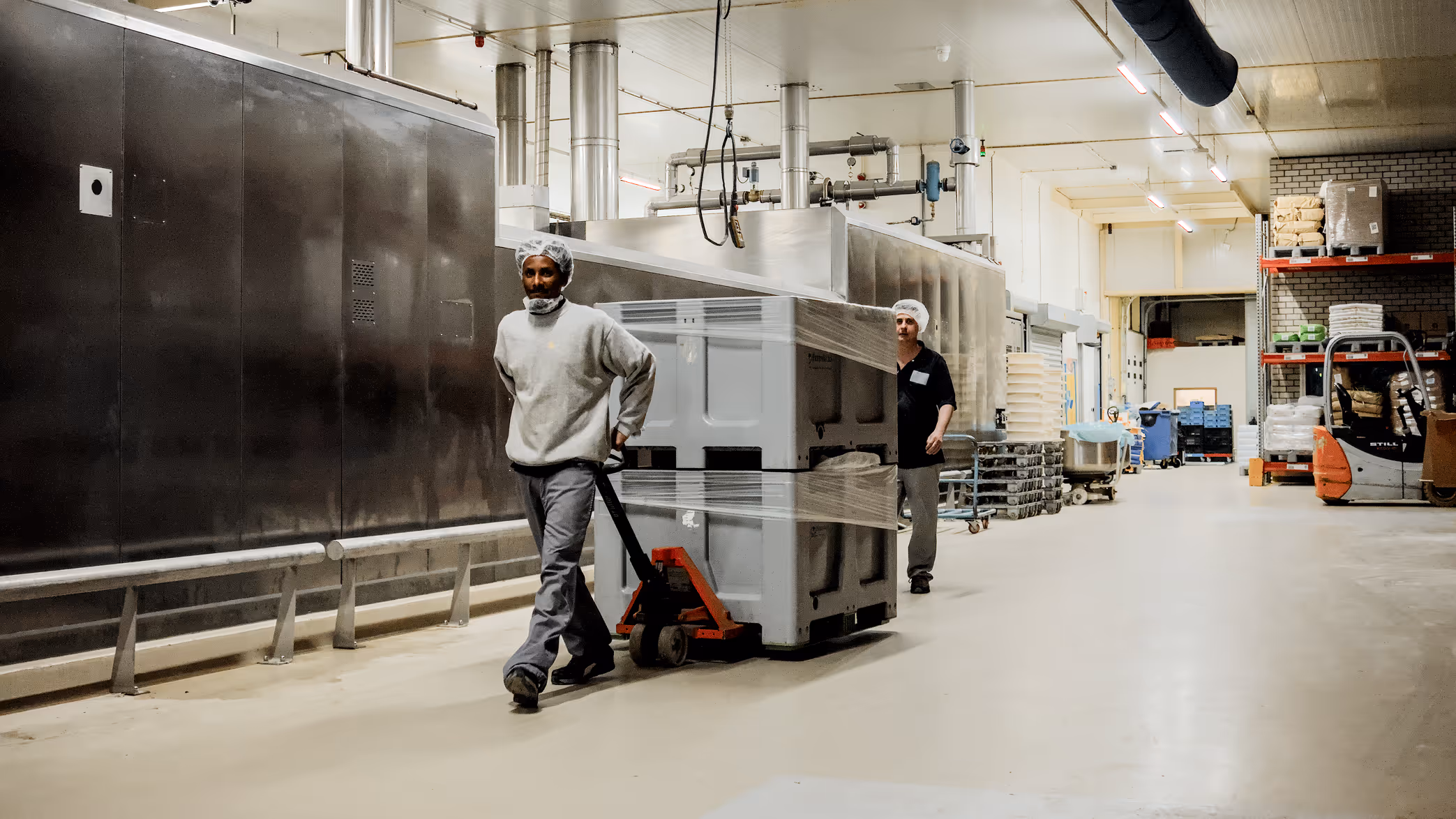 Twee arbeiders met haarnetjes vervoeren een grote, met folie verpakte container met een palletwagen in een fabriekshal.
