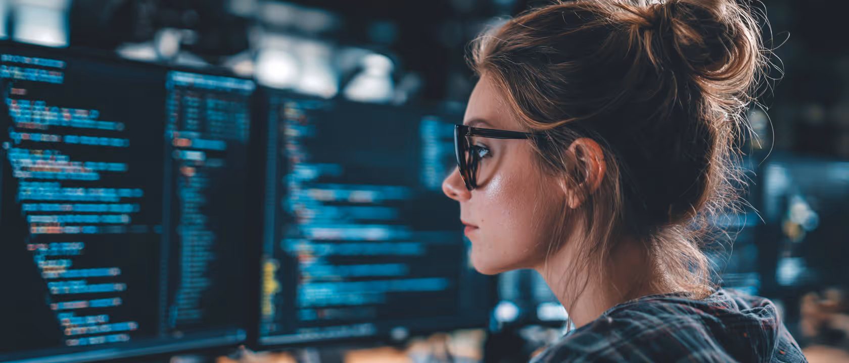 Femme concentrée portant des lunettes, regardant plusieurs écrans affichant du code informatique.