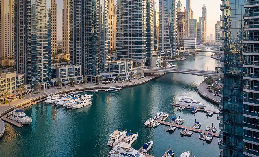 Marina with yachts docked alongside high-rise modern buildings and a bridge over calm water in a cityscape.