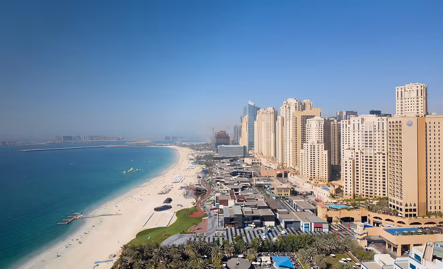 Aerial view of a long sandy beach with turquoise water beside a city skyline of tall beige buildings under a clear blue sky.