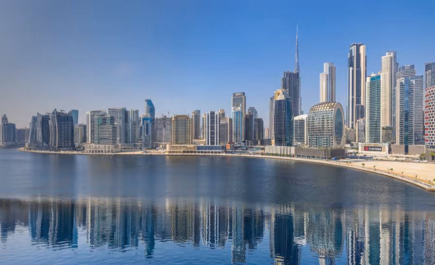 Panoramic view of modern skyscrapers along a waterfront with their reflections in the calm water under a clear blue sky.