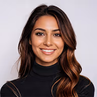 Smiling woman with long wavy brown hair wearing a black turtleneck against a light gray background.