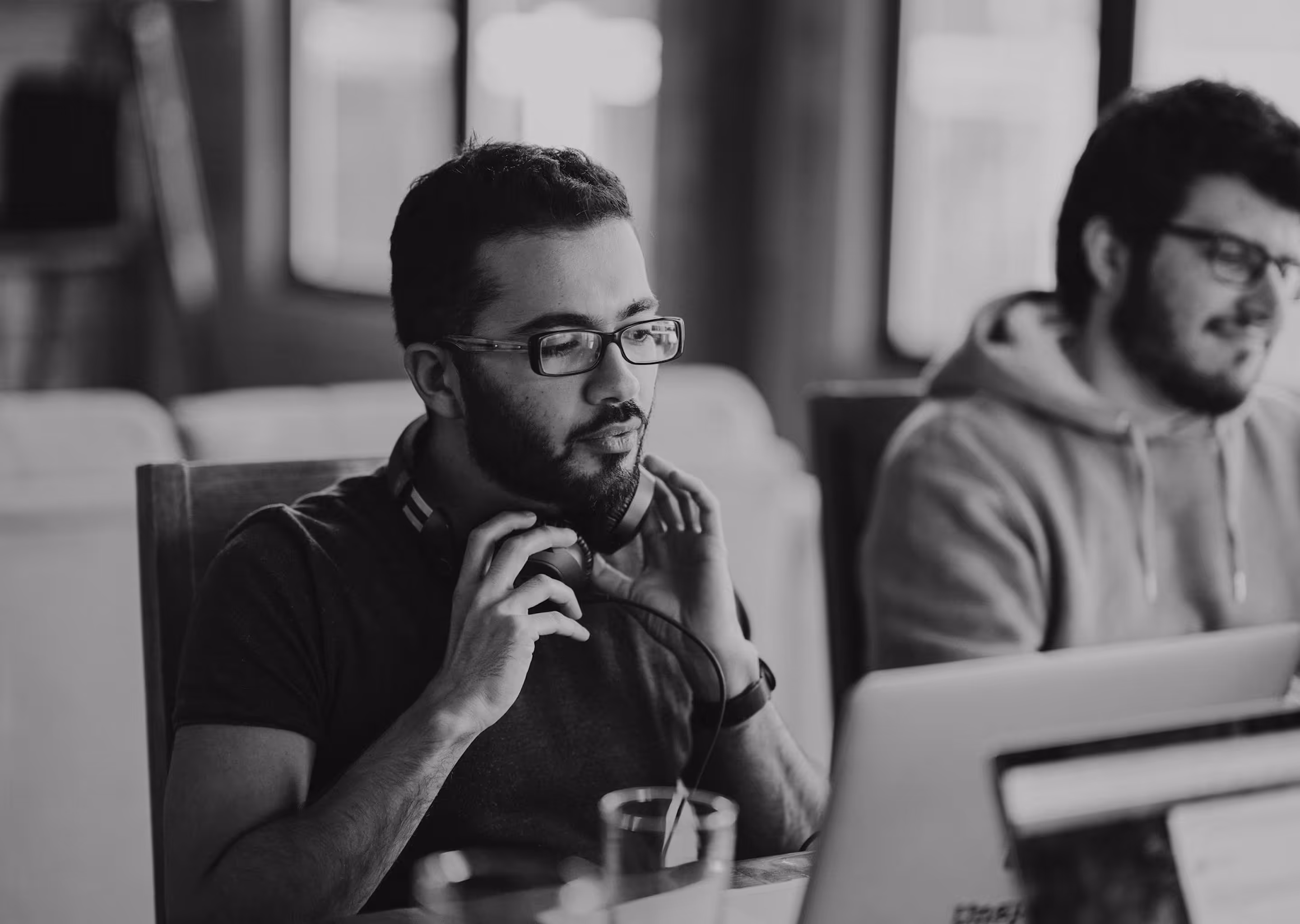 Two men wearing glasses are working together in an office, one with headphones around his neck in front of a laptop.