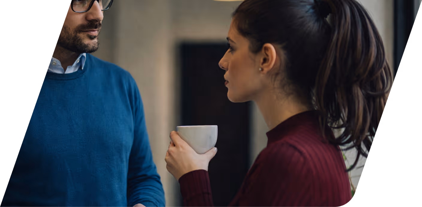 A woman holding a white cup looks at a man wearing a blue sweater and glasses during an indoor conversation.