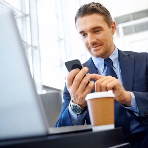 Man in a blue suit using a smartphone with a laptop and a coffee cup on a table.
