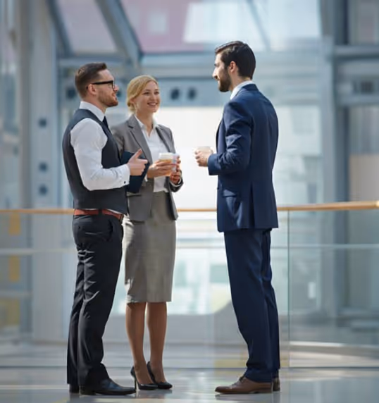 Three professionals in business clothes are talking while standing in a bright hall, holding coffee cups.
