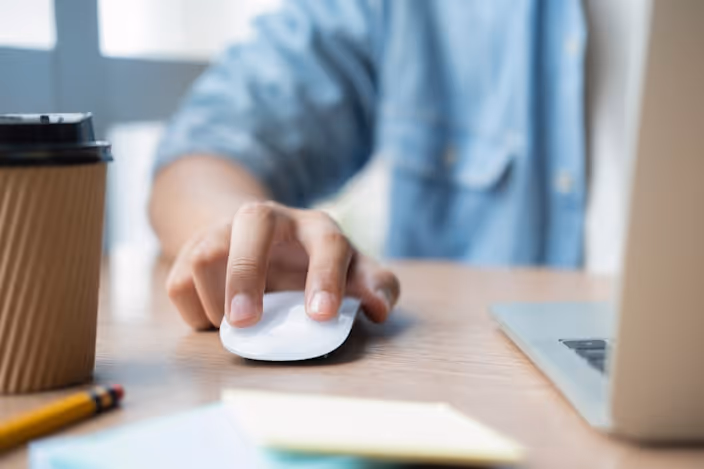 A hand using a computer mouse on a desk with a laptop, a coffee cup, and a pencil next to it.