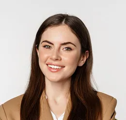 Smiling young woman with long brown hair wearing a tan blazer and white shirt, against a plain light background.