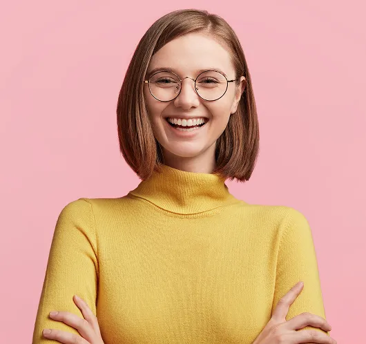 Smiling young woman with straight brown hair, round glasses, and yellow turtleneck sweater against a pink background.