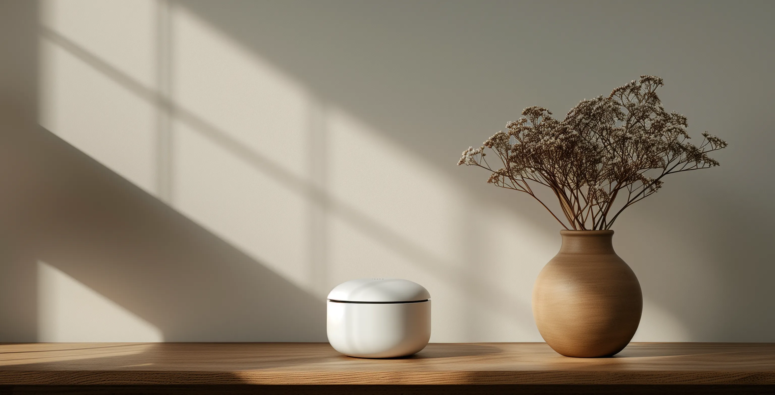 White round earbud case next to a wooden vase with dried flowers on a wooden table against a light wall with window shadows.