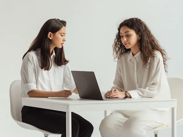Two women sitting at a white table, looking at a laptop in a bright room.