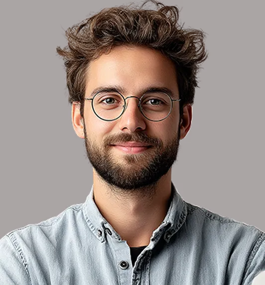 Smiling young man with curly hair, beard, and round glasses wearing a light denim shirt against a gray background.