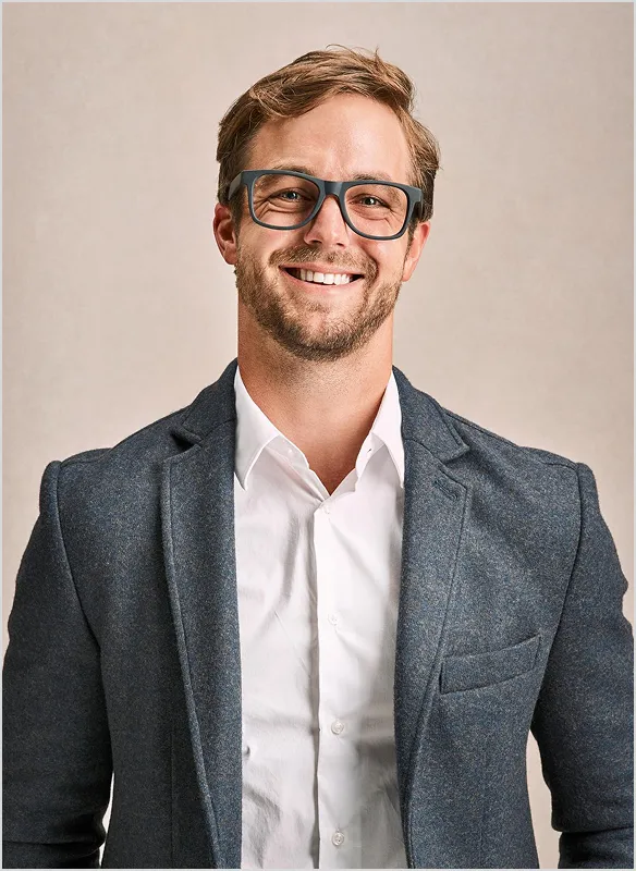Smiling man with beard and glasses wearing a gray blazer over a white shirt against a neutral background.