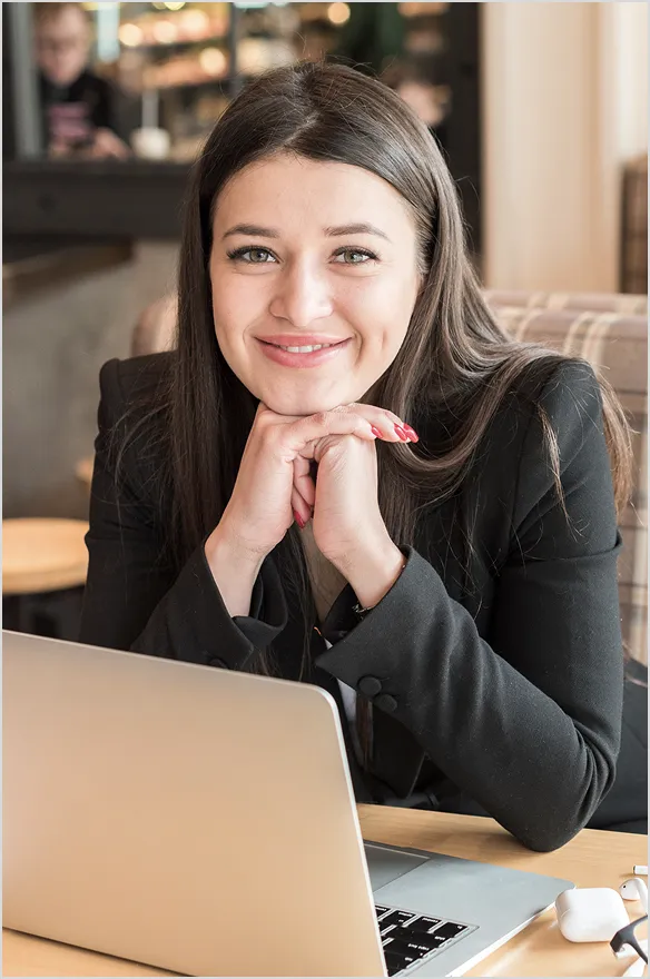Smiling woman with long dark hair resting her chin on folded hands in front of a laptop.