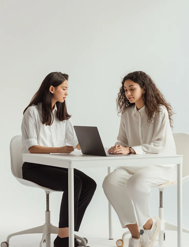 Two women in white shirts sitting at a white desk looking at a laptop together.