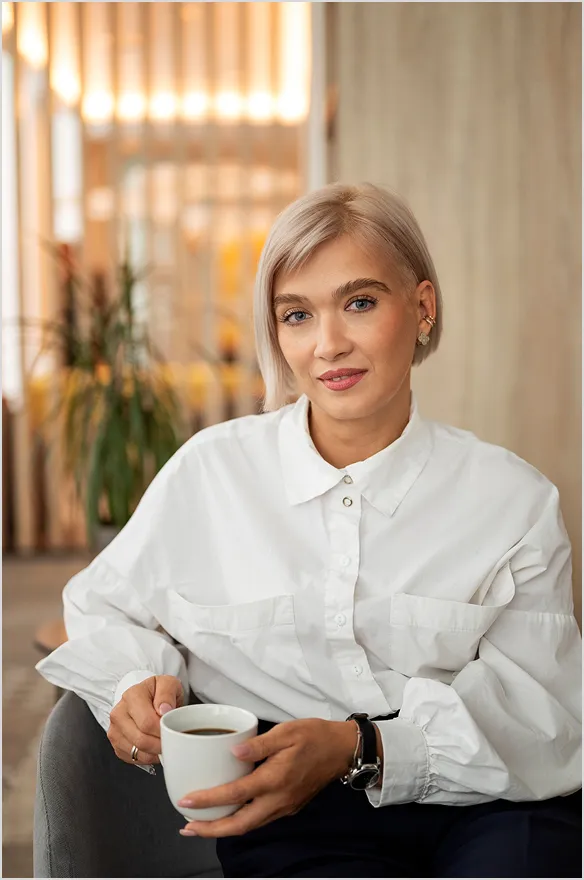 Woman with short blonde hair wearing a white shirt sitting on a chair and holding a white coffee cup.
