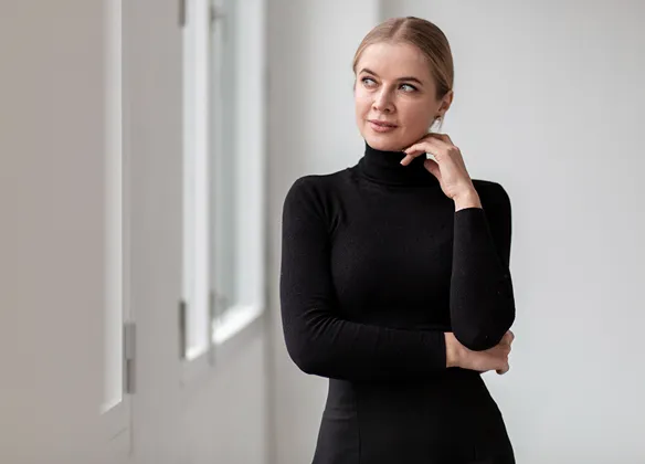 Woman in a black turtleneck standing thoughtfully by a window with white frames and a light gray wall background.