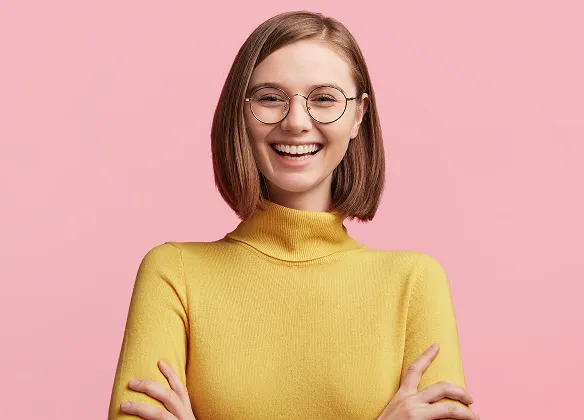 Smiling young woman with glasses wearing a yellow turtleneck sweater against a pink background.