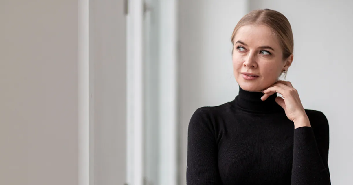 Blonde woman in black turtleneck looking thoughtfully to the side with hand near neck in a bright room.