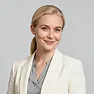 Smiling woman with blonde hair wearing a white blazer and light gray blouse against a gray background.