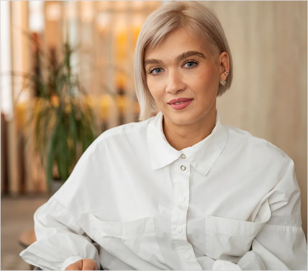 Smiling woman with short blonde hair wearing a white button-up shirt in a softly lit indoor setting.
