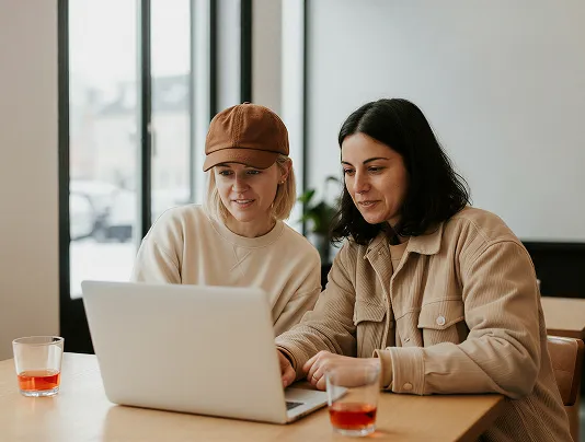 Two women sitting at a table looking at a laptop, with glasses of red drink nearby.