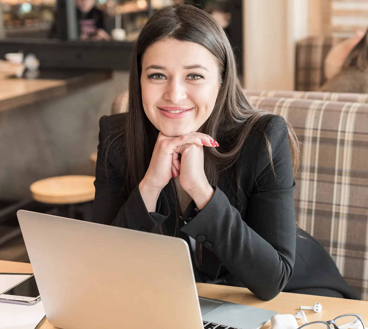 Smiling woman with long brown hair sitting at a table with a laptop in a cozy café.