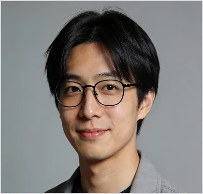 Portrait of a young man with black hair, wearing black-rimmed glasses and a light gray jacket, smiling gently against a neutral gray background.
