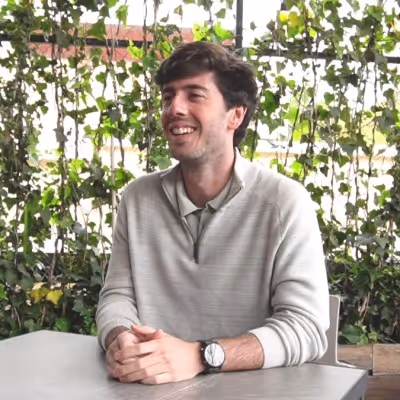 Hombre sonriente con suéter gris sentado en una mesa al aire libre con plantas verdes de fondo.
