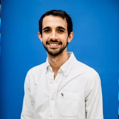 Hombre joven sonriente con barba y camisa blanca frente a fondo azul.
