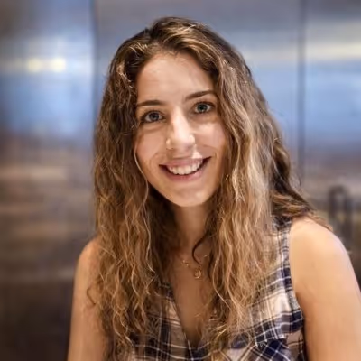 Mujer sonriente con cabello rizado y ropa de cuadros, en un interior con fondo desenfocado.