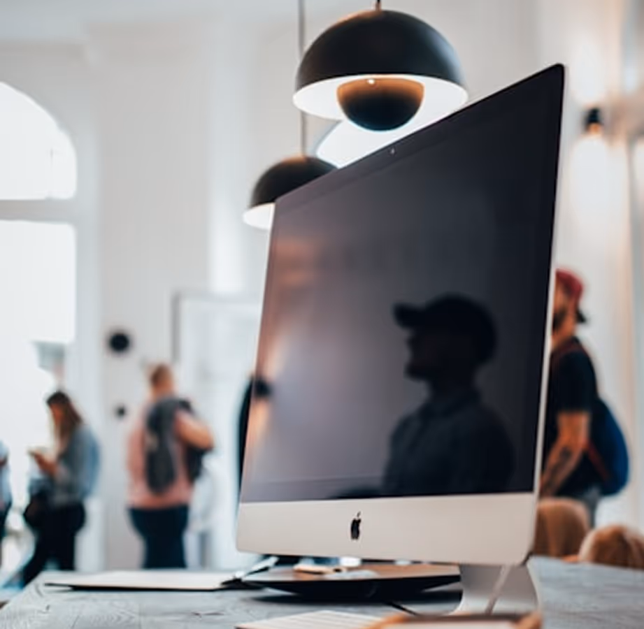 Pantalla de computadora iMac apagada en un escritorio con personas desenfocadas en el fondo de una oficina luminosa.