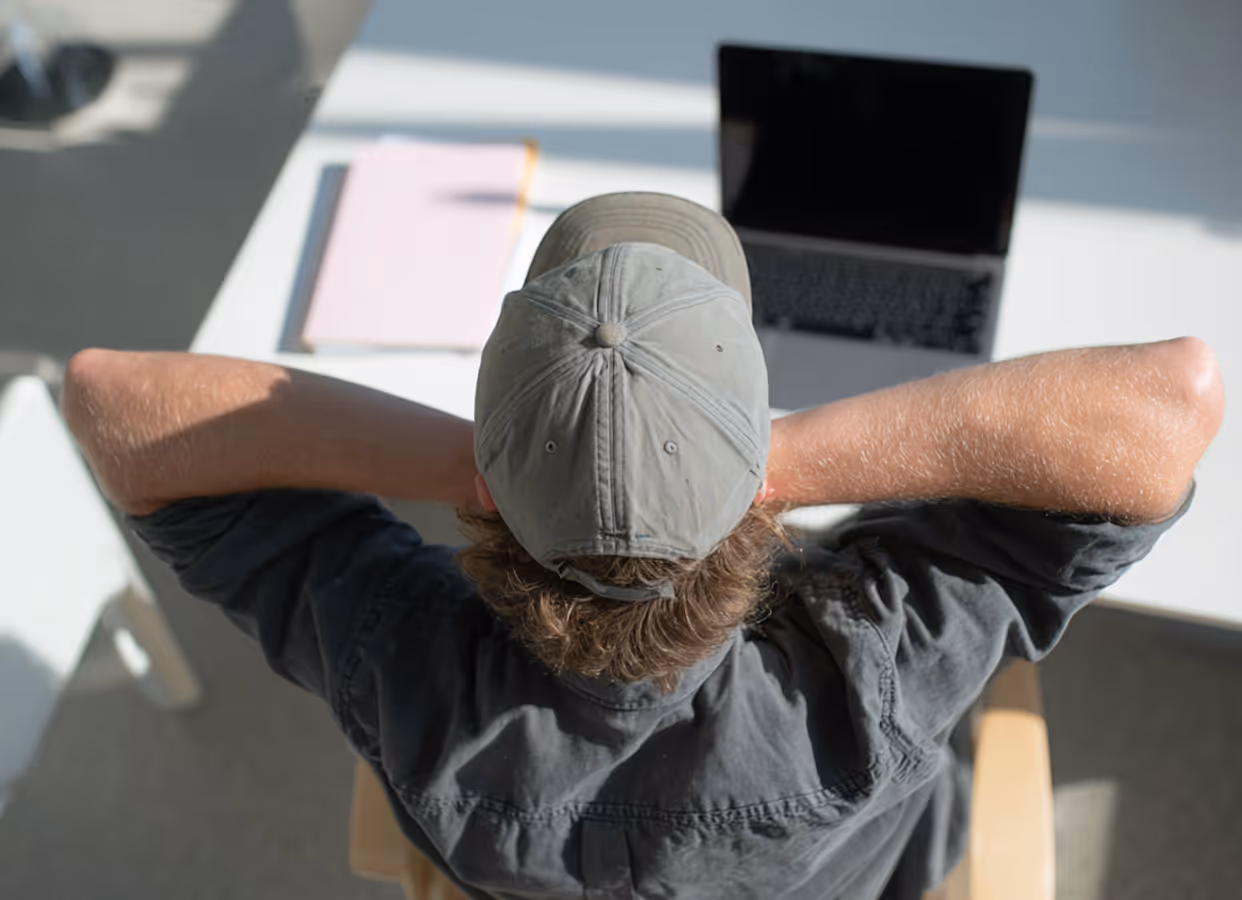 Persona con gorra gris y camisa oscura recostada en silla frente a una mesa blanca con computadora portátil y cuaderno.