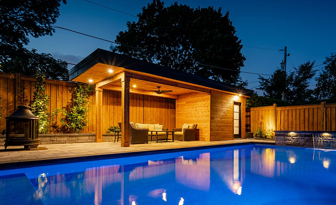 Lit wooden poolside cabana with seating area next to a blue illuminated swimming pool at dusk.