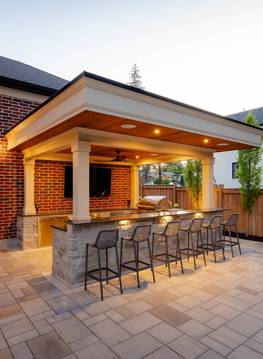 Outdoor covered barbecue area with stone bar counter, six black bar stools, built-in grill, ceiling fan, and mounted TV on brick wall.
