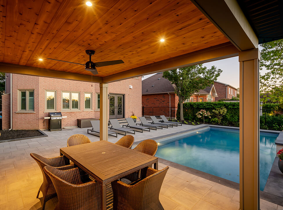 Covered patio with wooden ceiling, ceiling fan, wicker dining set, and outdoor pool surrounded by lounge chairs and brick houses in the evening.