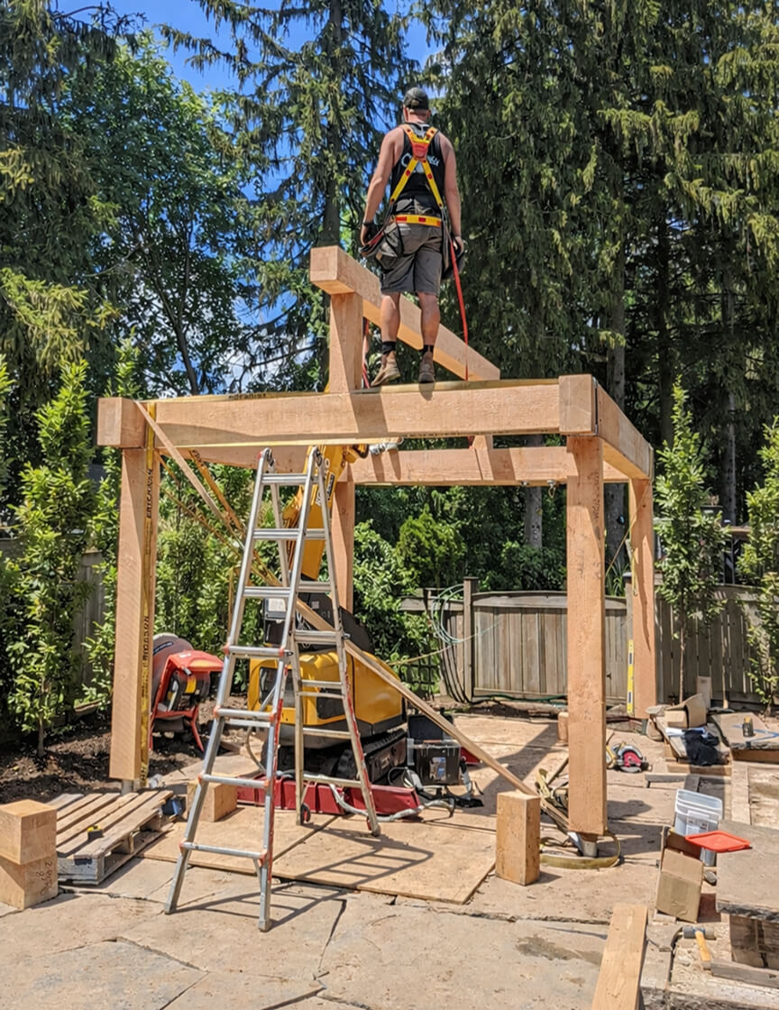 Construction worker wearing safety harness standing on wooden beams building a wooden frame structure outdoors with tools and ladder nearby.