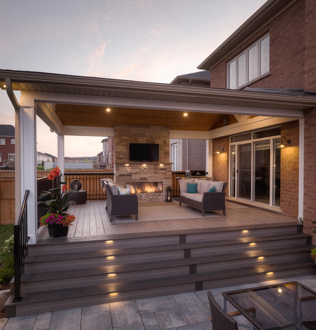 Covered outdoor patio at dusk with two sofas facing a stone fireplace with TV mounted above.