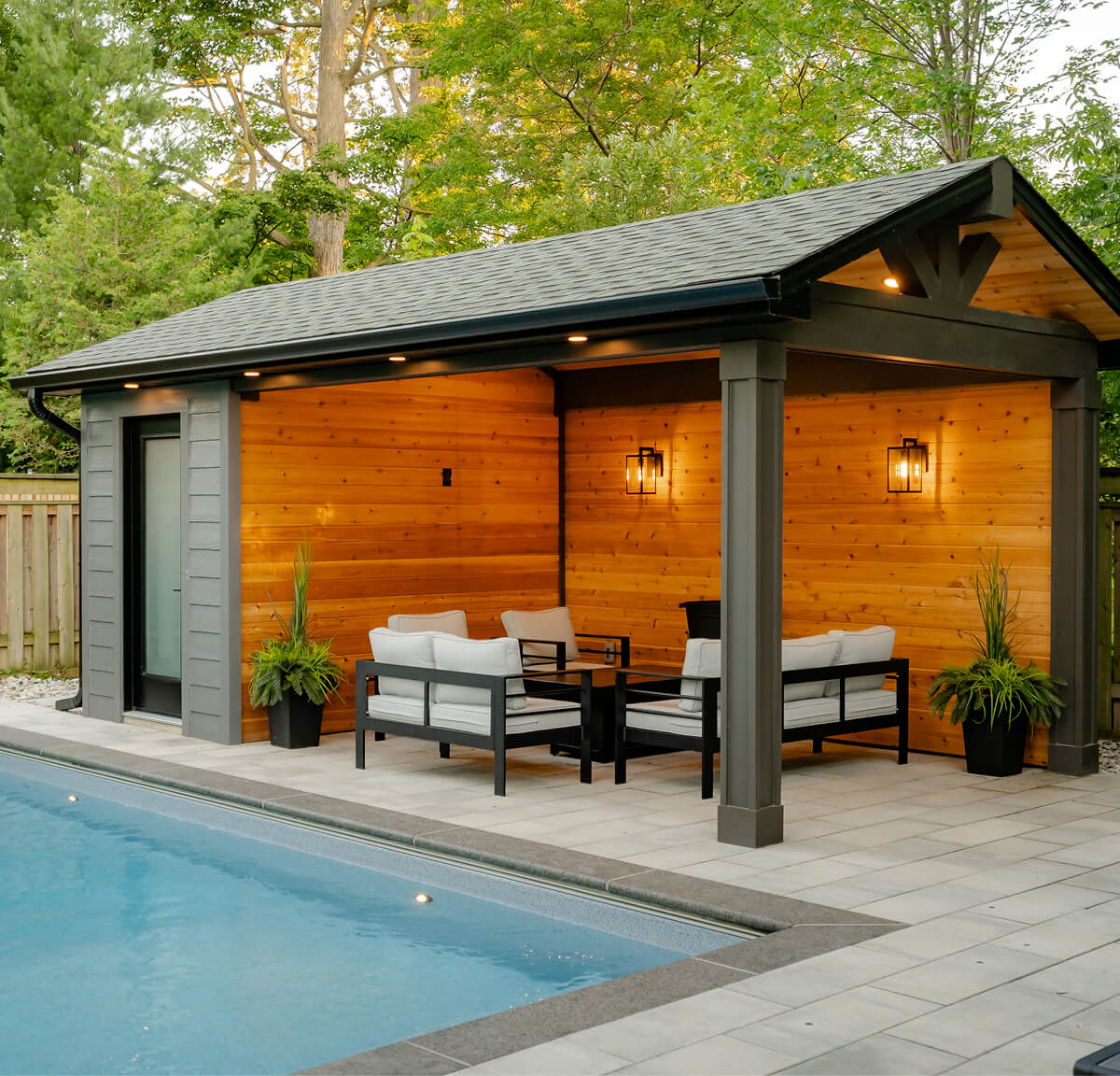 Covered patio with wooden walls, modern outdoor seating and plants beside a blue swimming pool.