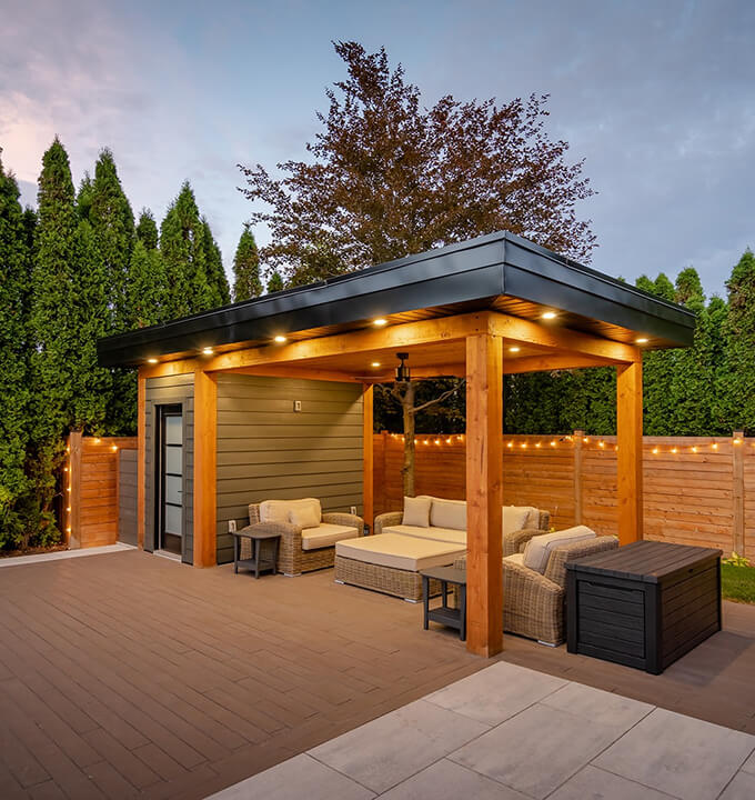 Modern outdoor patio with wooden pergola, wicker furniture, and string lights along wooden fence at dusk.