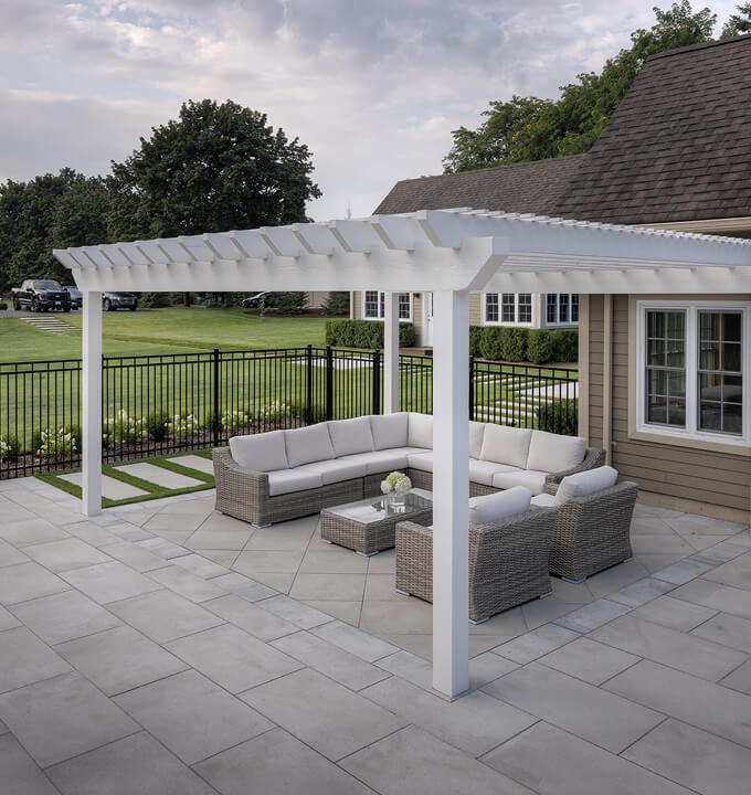 Outdoor patio with white pergola covering gray cushioned wicker sectional sofa and two chairs on large stone tiles beside house.