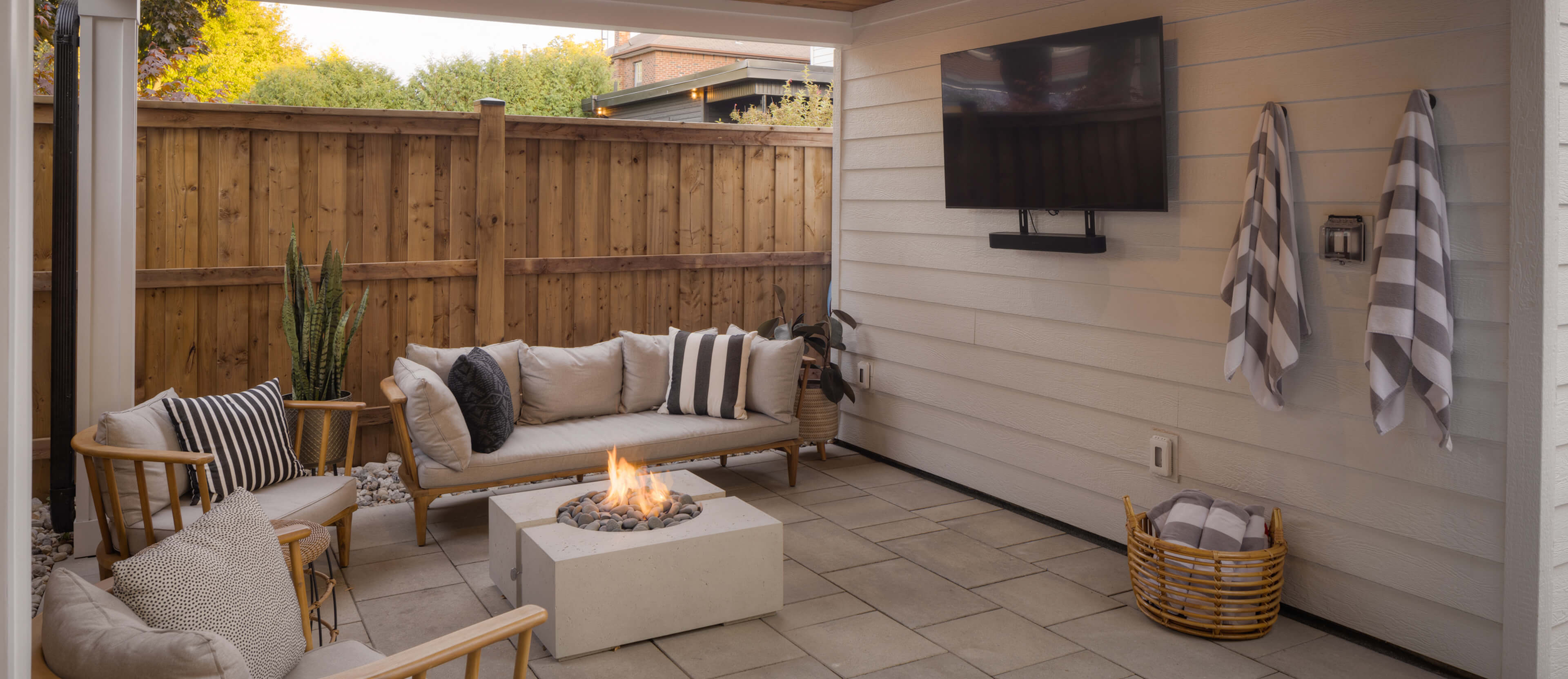 Cozy outdoor patio with cushioned wooden seating, a modern fire pit, wall-mounted TV, and striped towels hanging on a white wall.