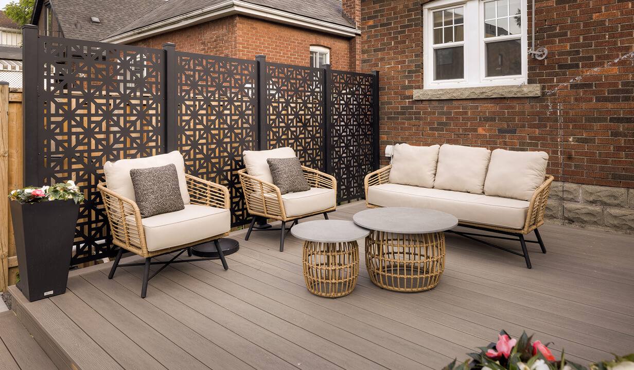 Modern outdoor patio set with beige cushioned wicker sofa and chairs, round coffee tables, and black geometric privacy screens against a brick house.