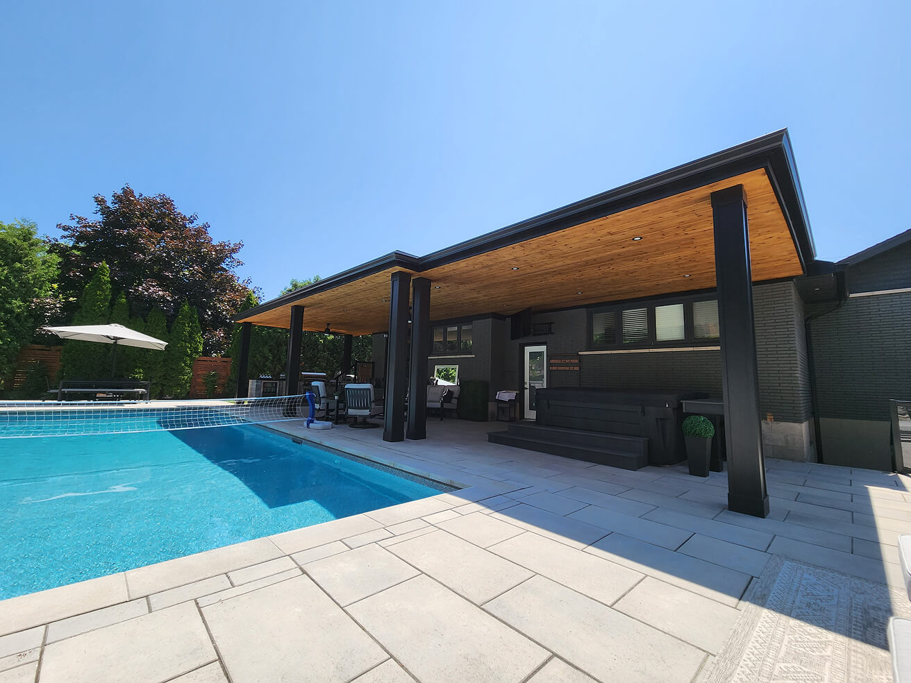 Modern outdoor patio area with a covered wooden ceiling, black pillars, seating, and a blue swimming pool with a volleyball net.