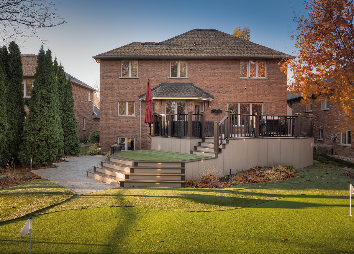 Backyard of a two-story brick house with a raised deck, stairs, and a small putting green in the foreground.